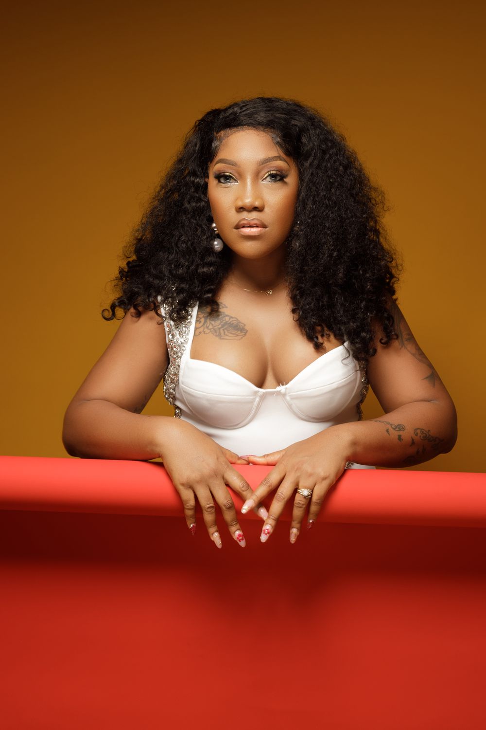 Fashion portrait against brown background showing curly hair and white lingerie top with hands resting on red surface.