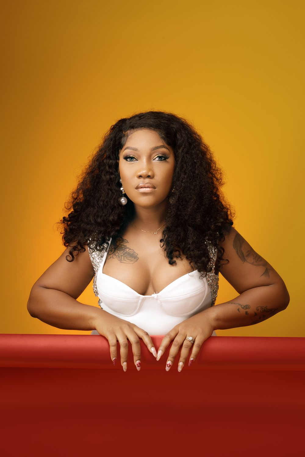 Studio portrait against orange background featuring curly hair, white top and red table with elegant jewelry and styling.