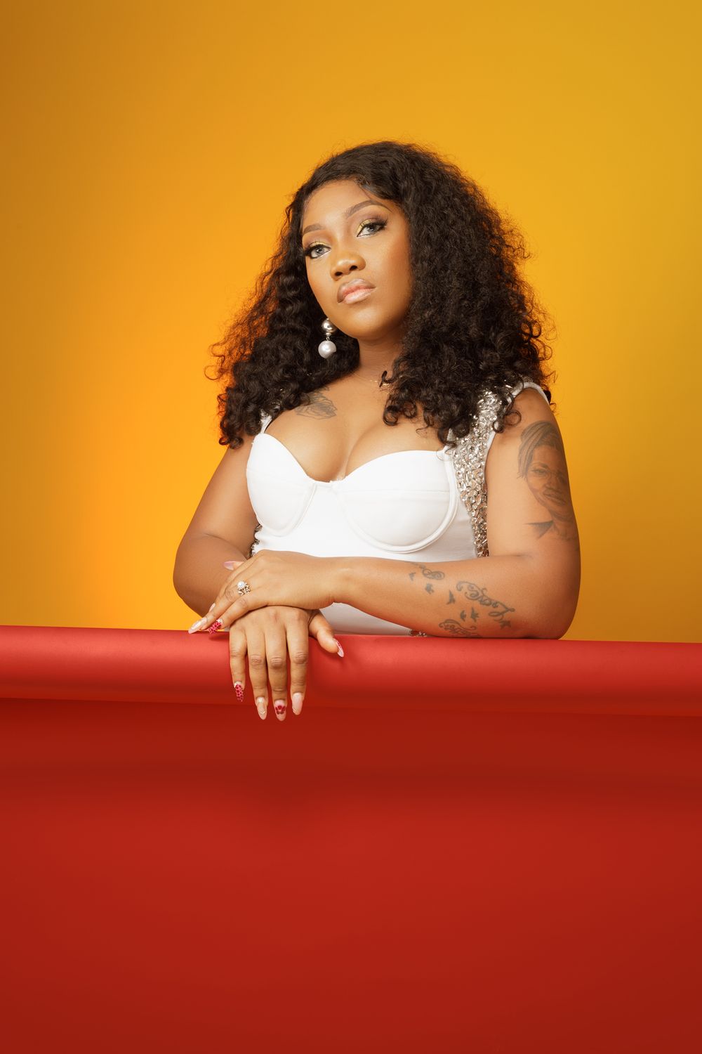 Model in white top with curly hair poses against vibrant orange backdrop and red table.