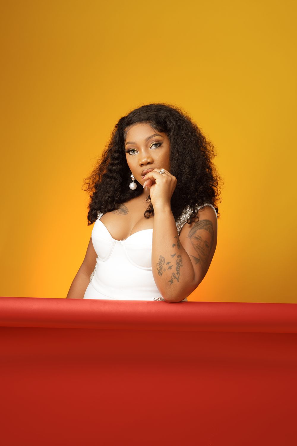 A person in a white top poses confidently against a vibrant orange background with a red surface in the foreground.