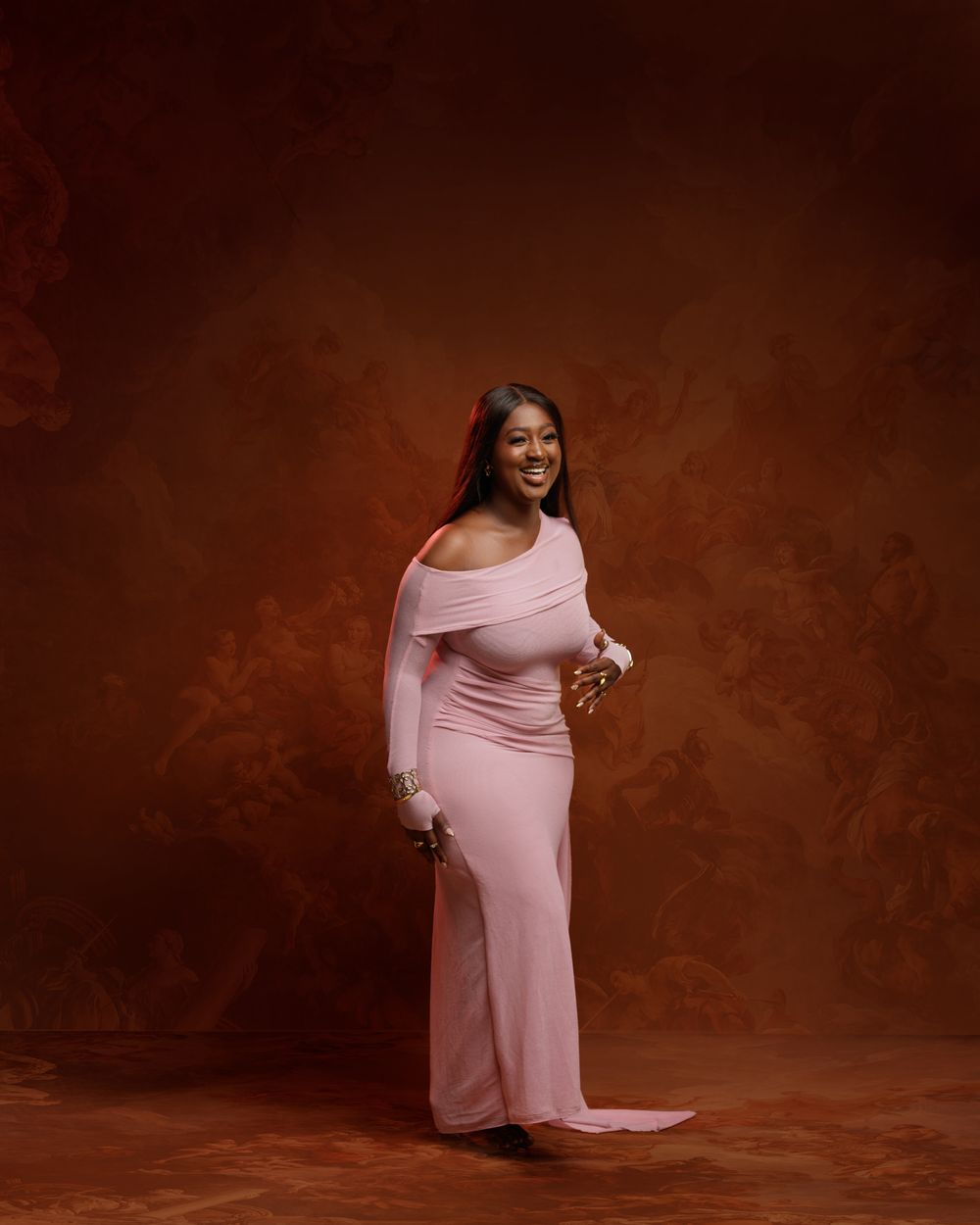 A person poses in a pink off-shoulder maternity gown against a brown backdrop during a professional photography session.