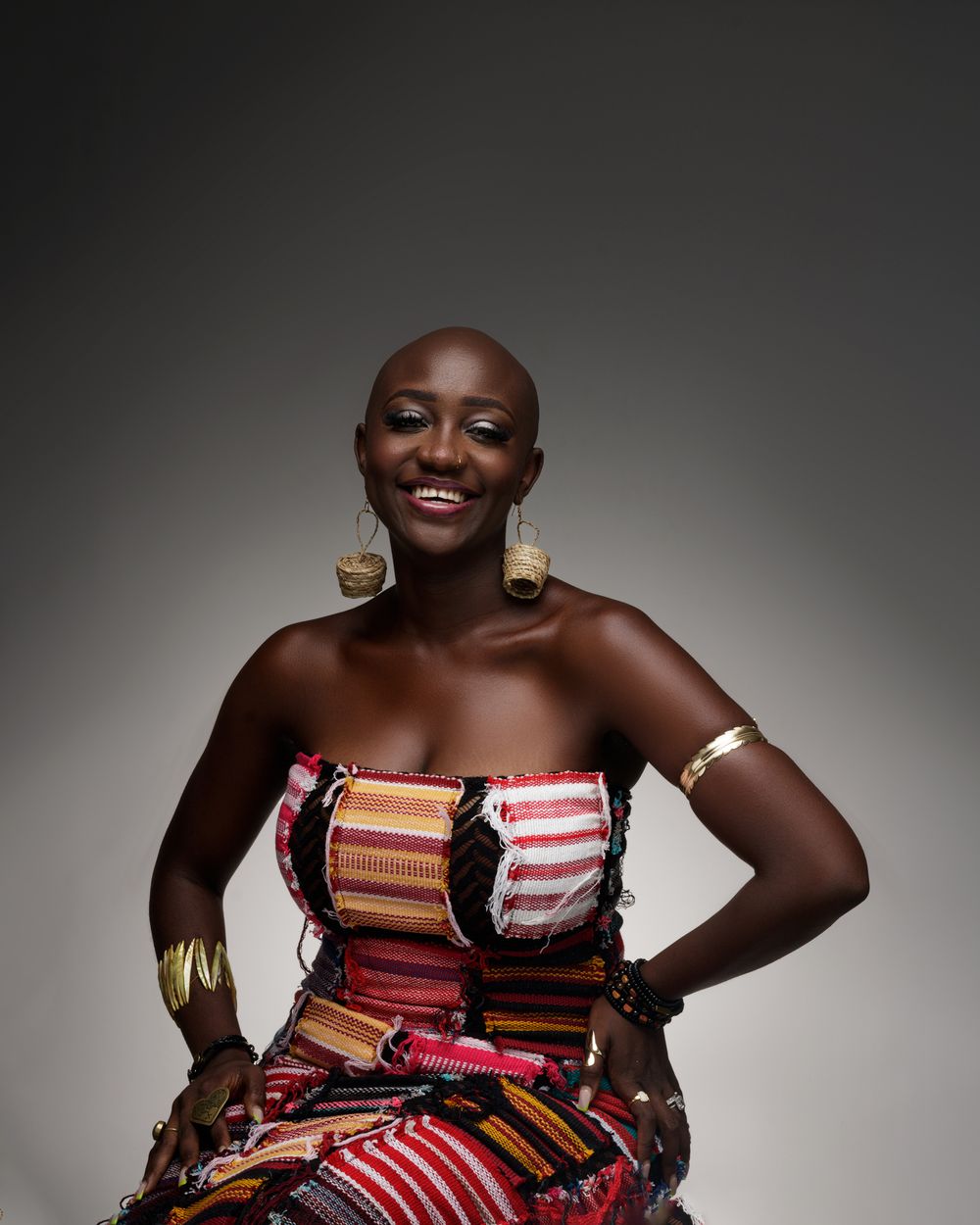 Smiling creative fashion portrait in a colorful striped strapless dress with gold statement jewelry against dark background.