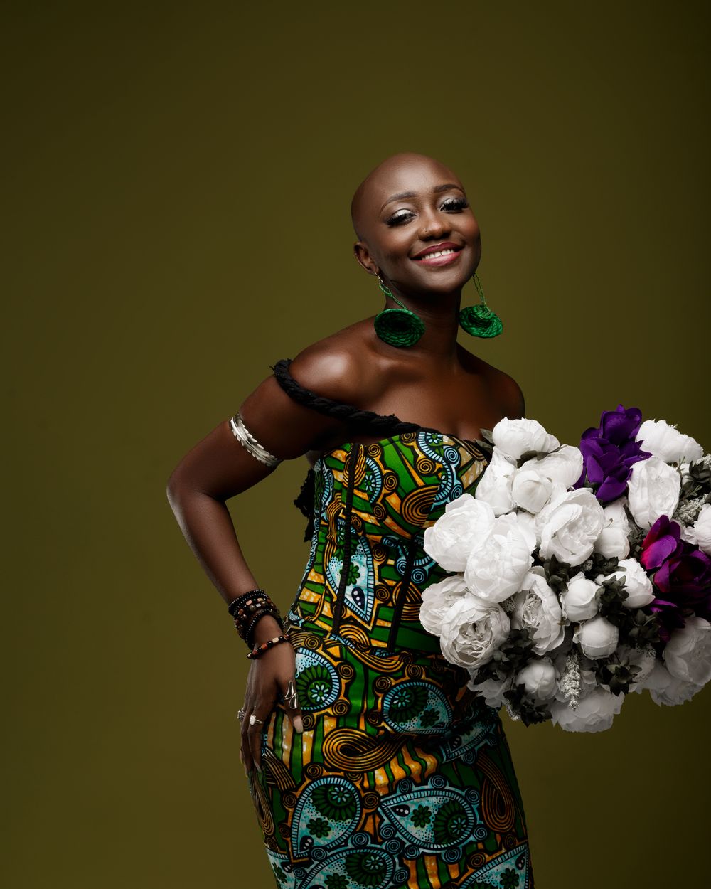 Elegant portrait against olive backdrop showing someone in vibrant African print dress holding white and purple flowers.