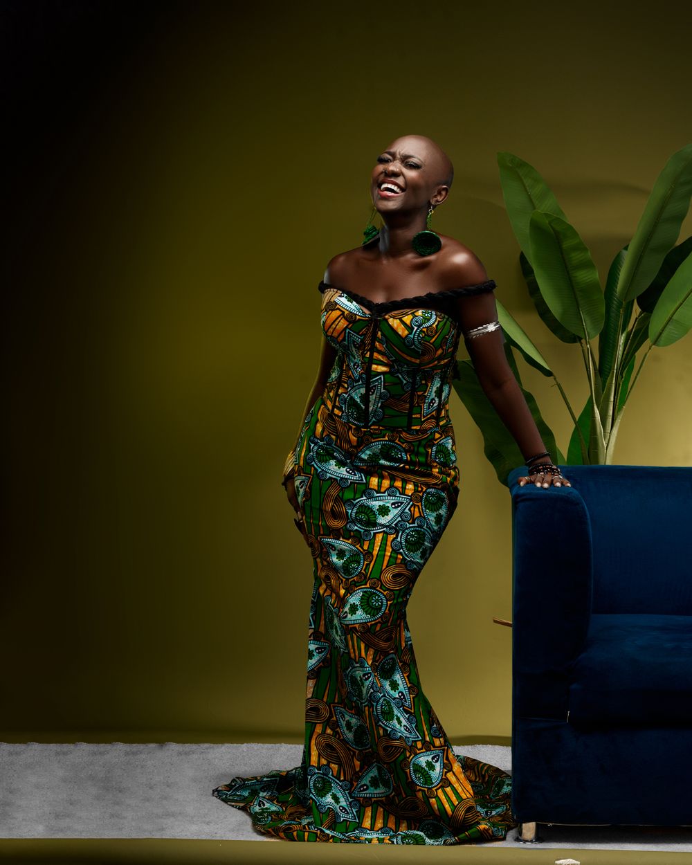 A figure in a vibrant patterned green and orange mermaid-style gown poses dramatically against an olive backdrop with potted plants.