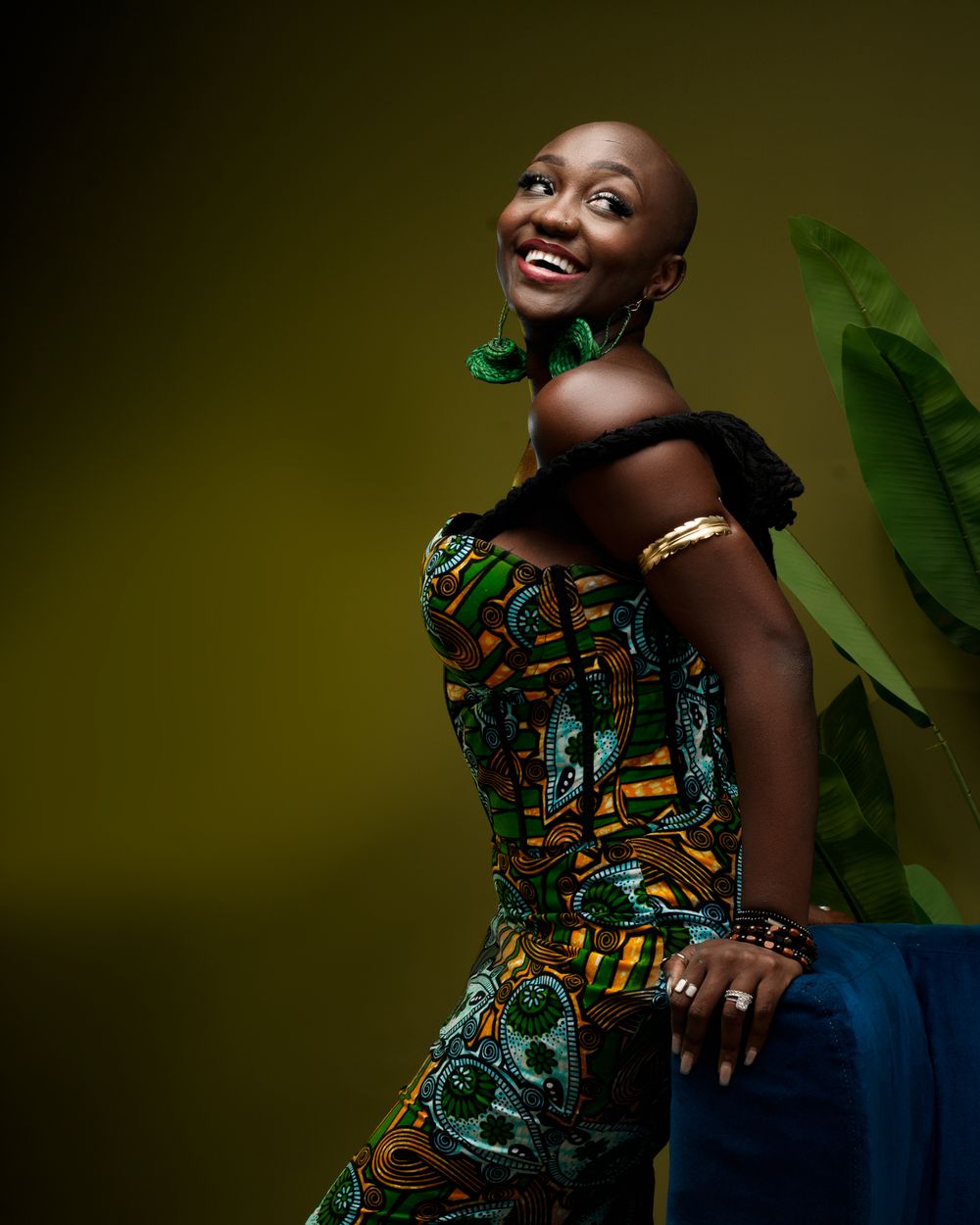 Joyful model in vibrant African print dress poses gracefully against dark green backdrop with tropical leaves.