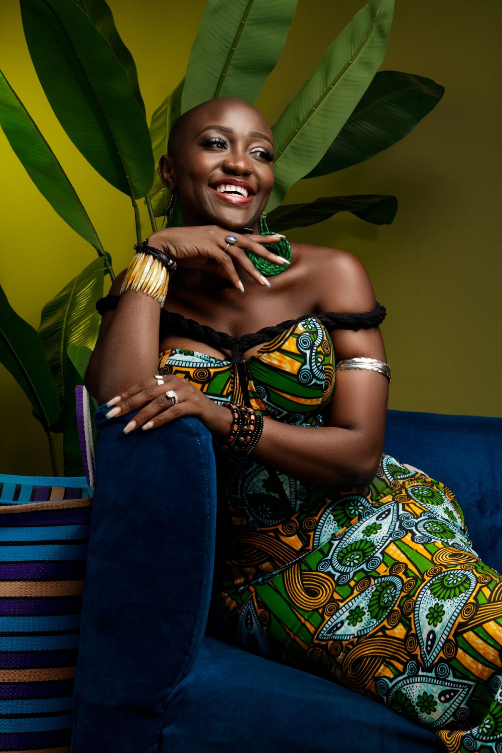 Fashion portrait of someone in vibrant African print dress sitting on blue sofa with tropical leaves against yellow backdrop.