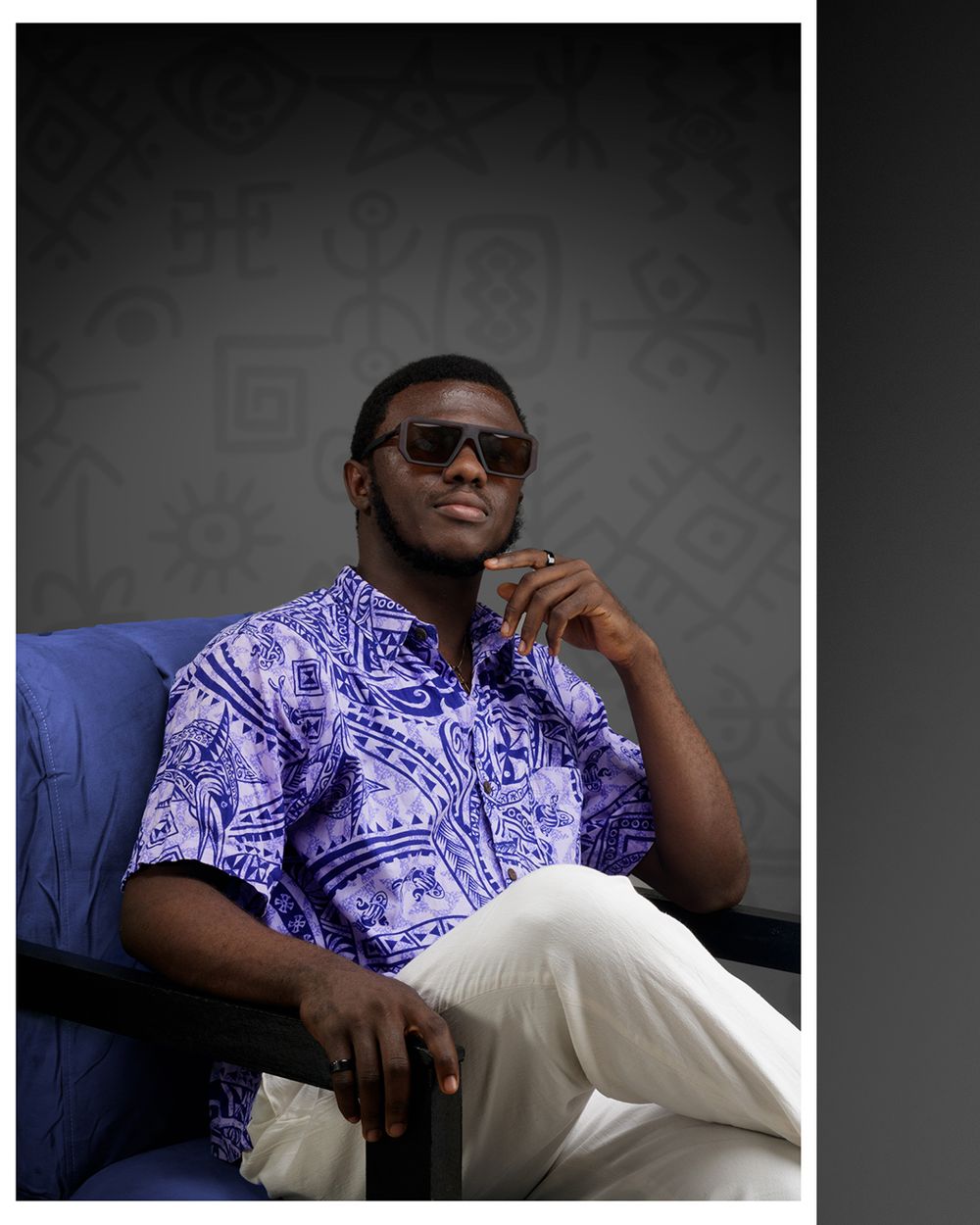 A person in sunglasses and a purple patterned shirt sits thoughtfully in a blue chair against a dark background.