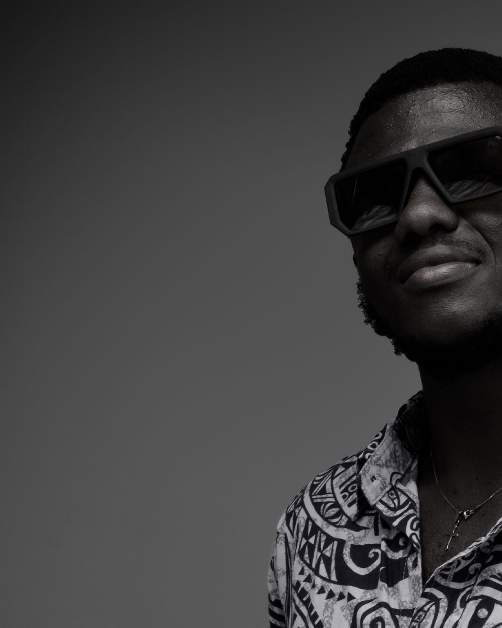 A person wearing sunglasses and a patterned shirt smiles in a black and white photograph against a plain backdrop.