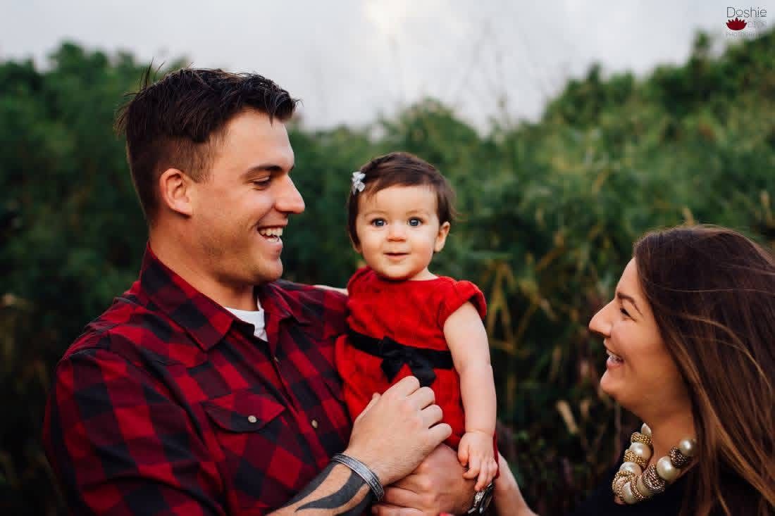 A happy young couple in red outfits smiling at their baby girl in a red dress outdoors.