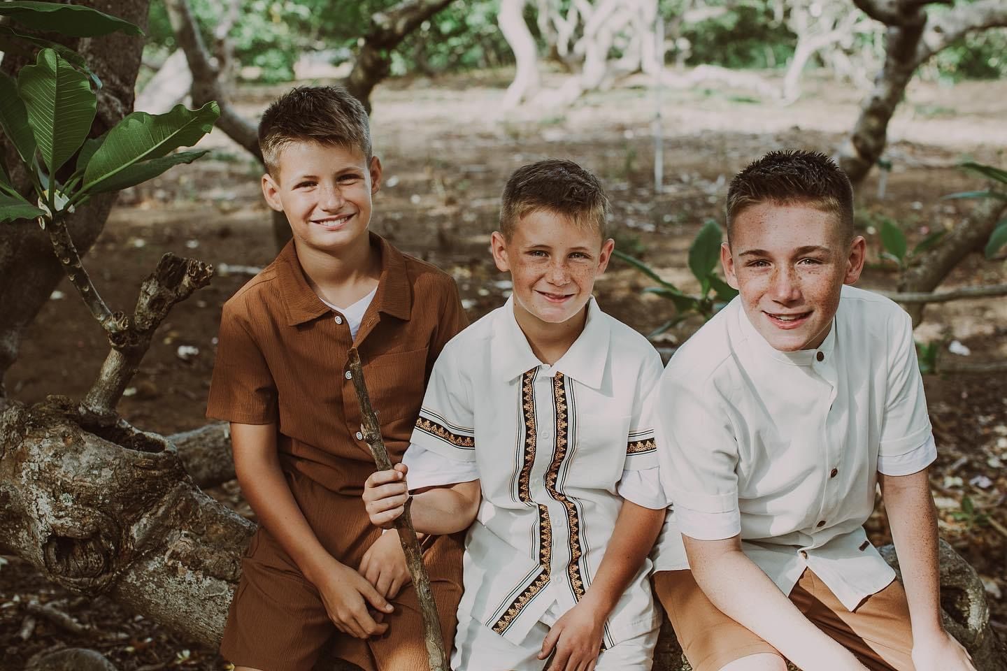 Three smiling boys sitting together on a tree branch outdoors in a lush green garden setting.
