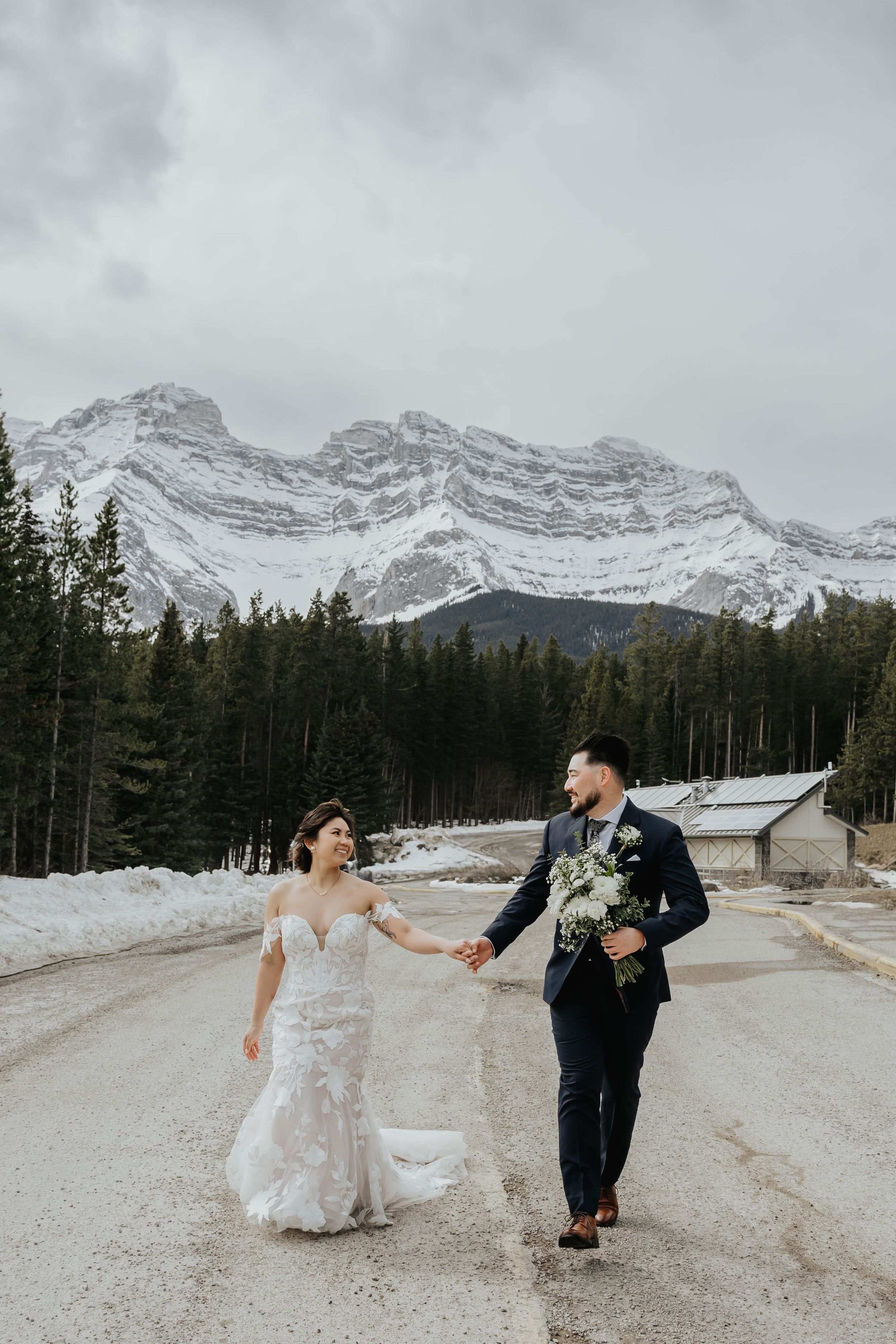 Wedding couple walking hand in hand with snow-capped mountain peaks in background.