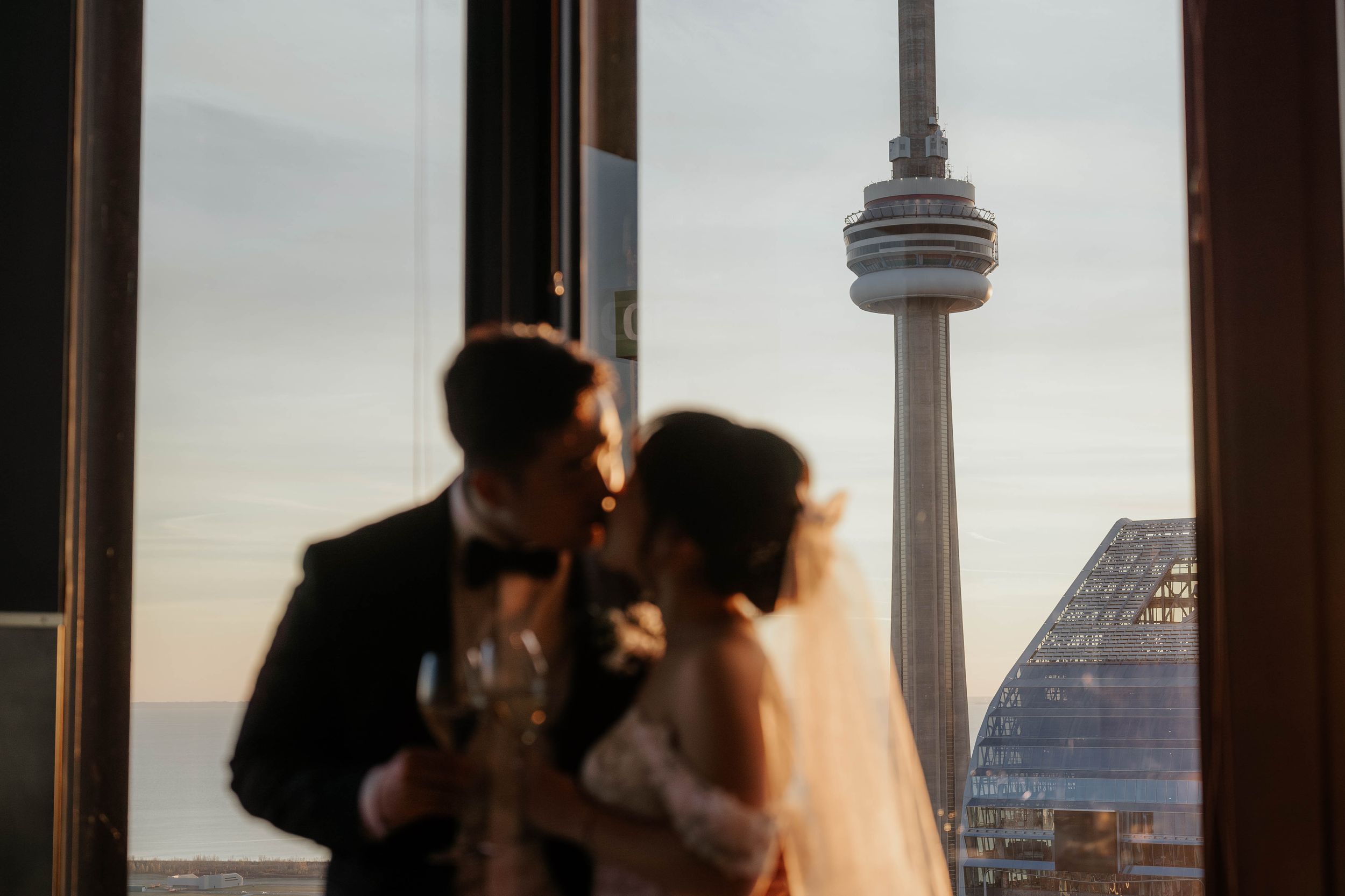 Couple sharing an intimate moment with Toronto's CN Tower visible through window at sunset.