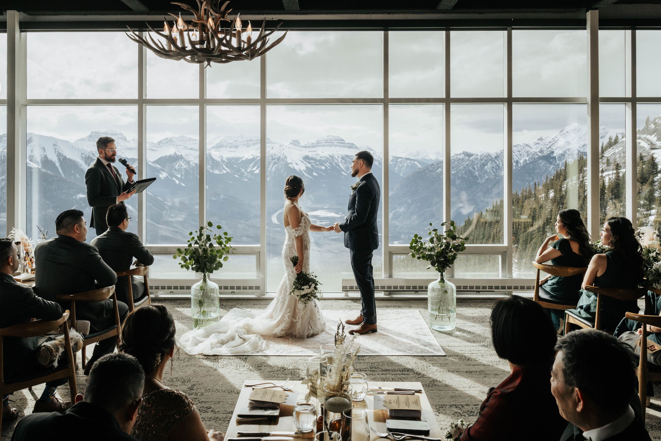 Intimate wedding ceremony taking place in front of floor-to-ceiling windows overlooking snowy mountain peaks.