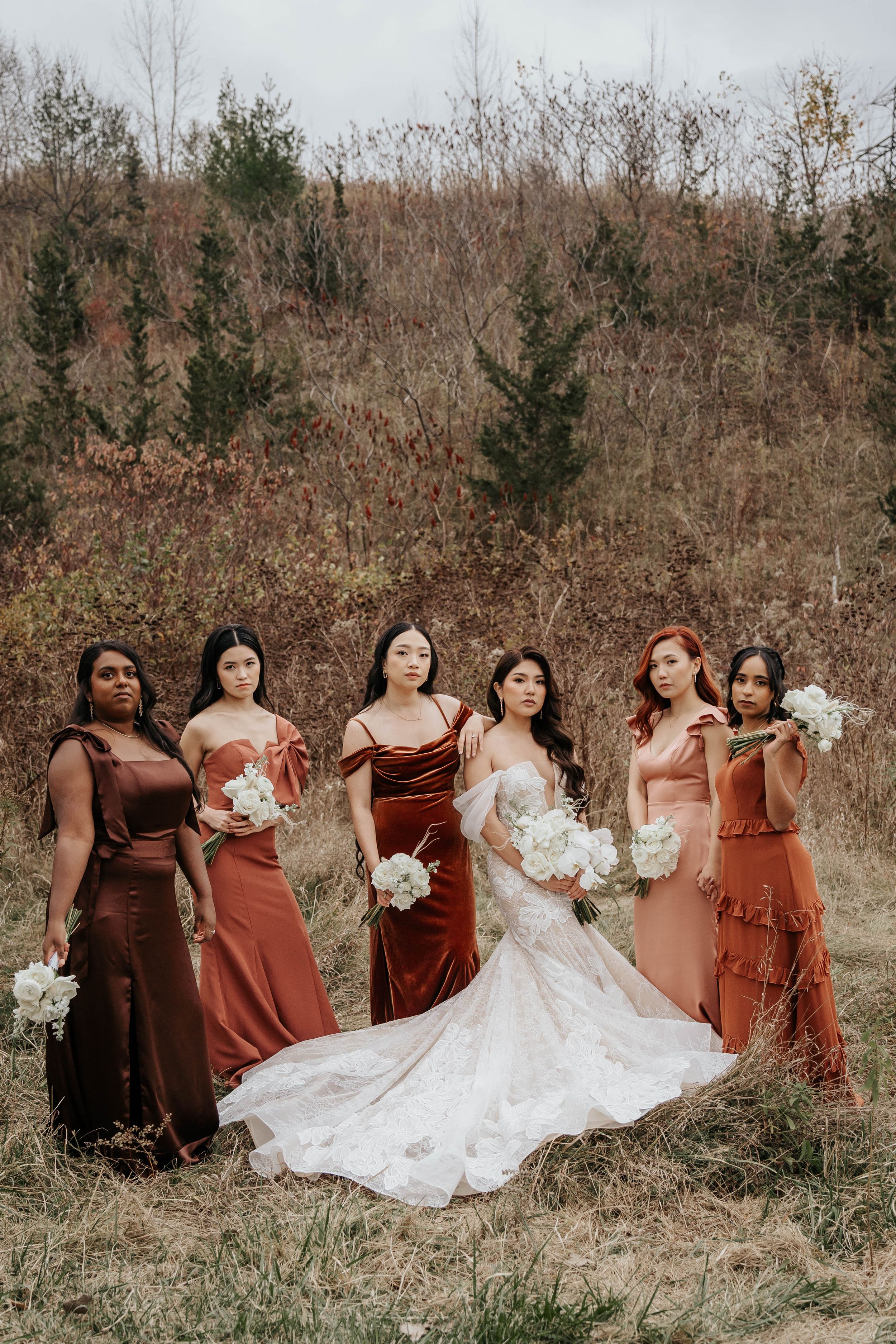 Bridal party in rust and burgundy dresses pose together in autumn field setting.