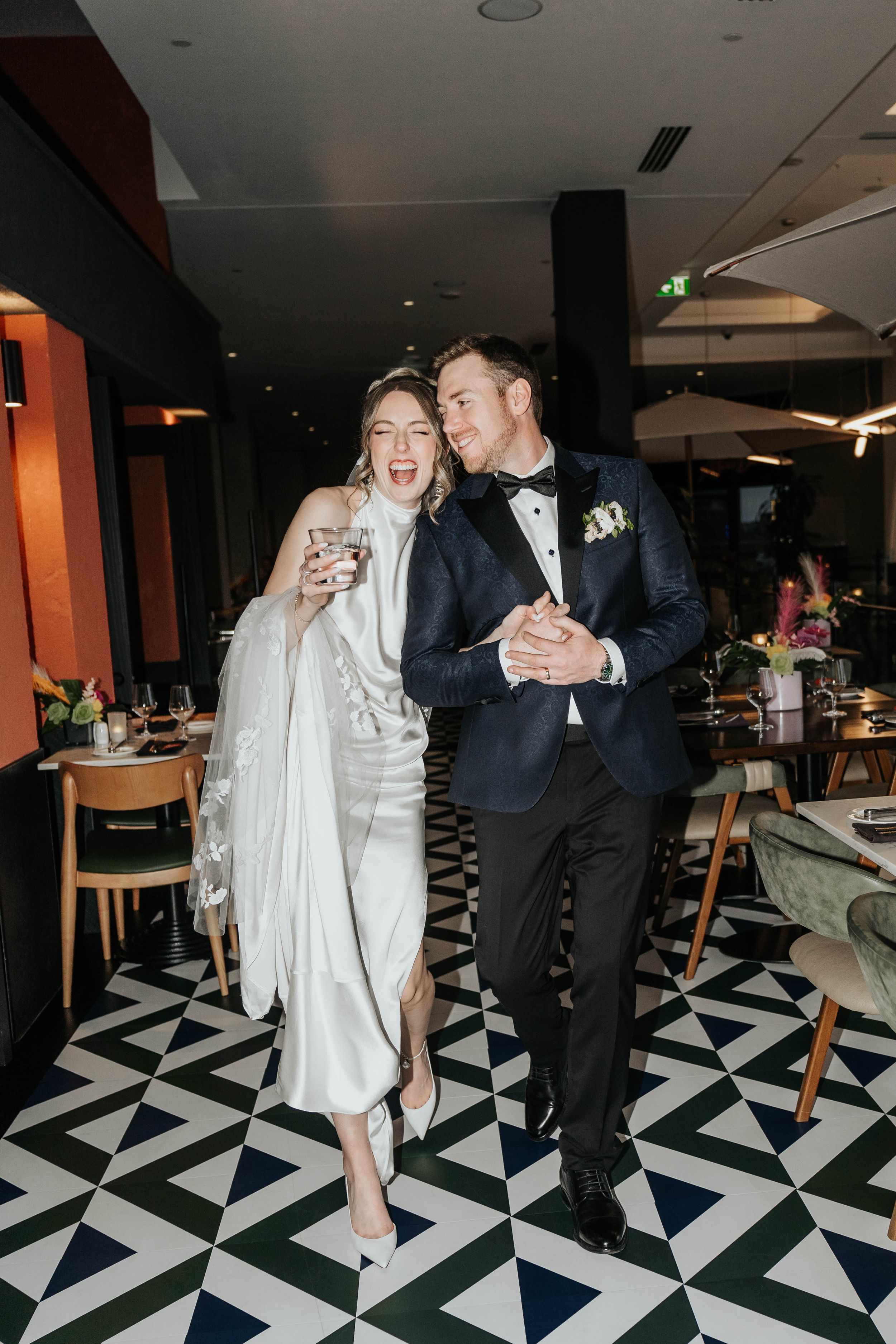 Newlyweds share drinks while walking across geometric patterned floor at reception venue.