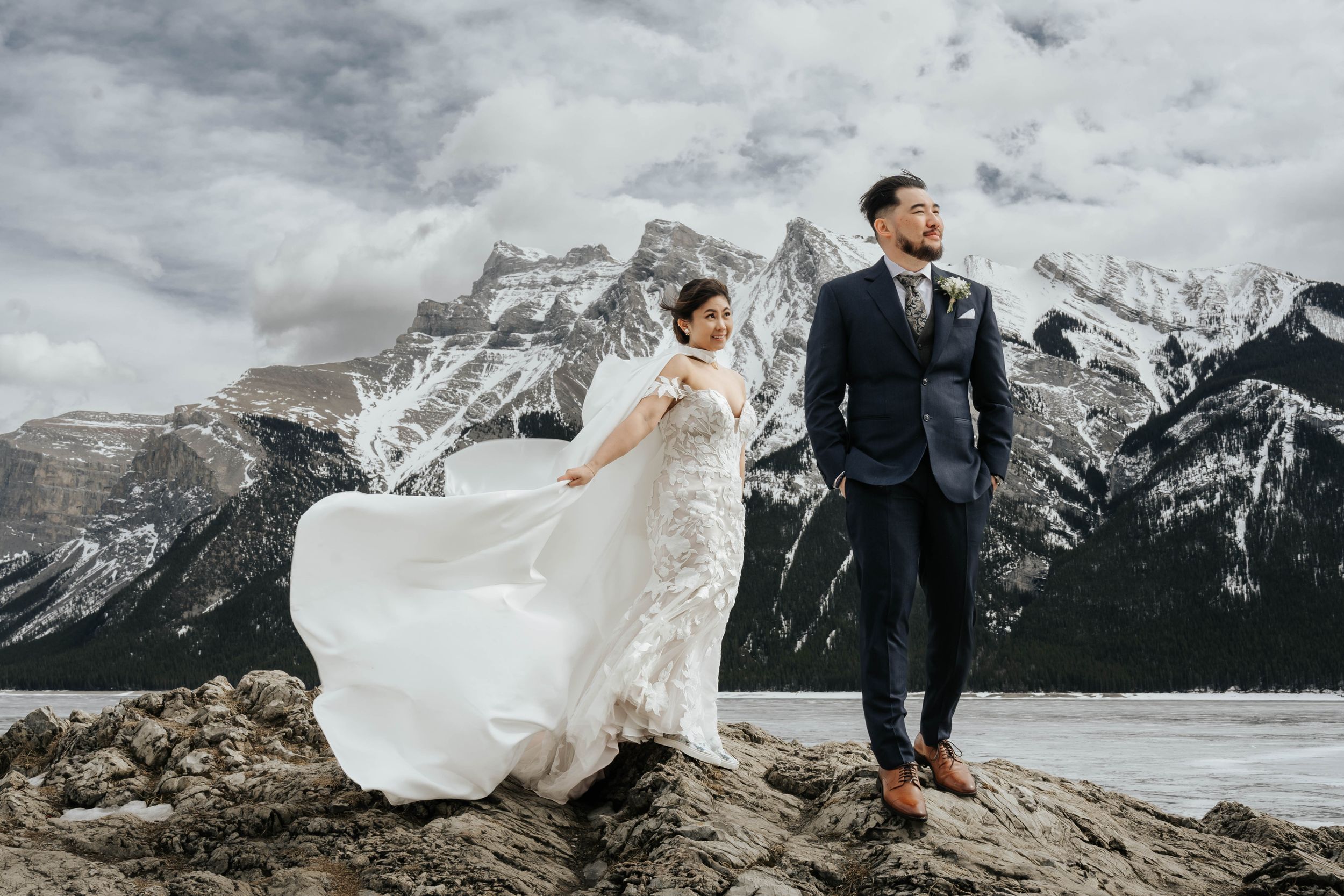 Bride's dress flowing in wind as couple pose dramatically on mountain peak with rocky landscape backdrop.