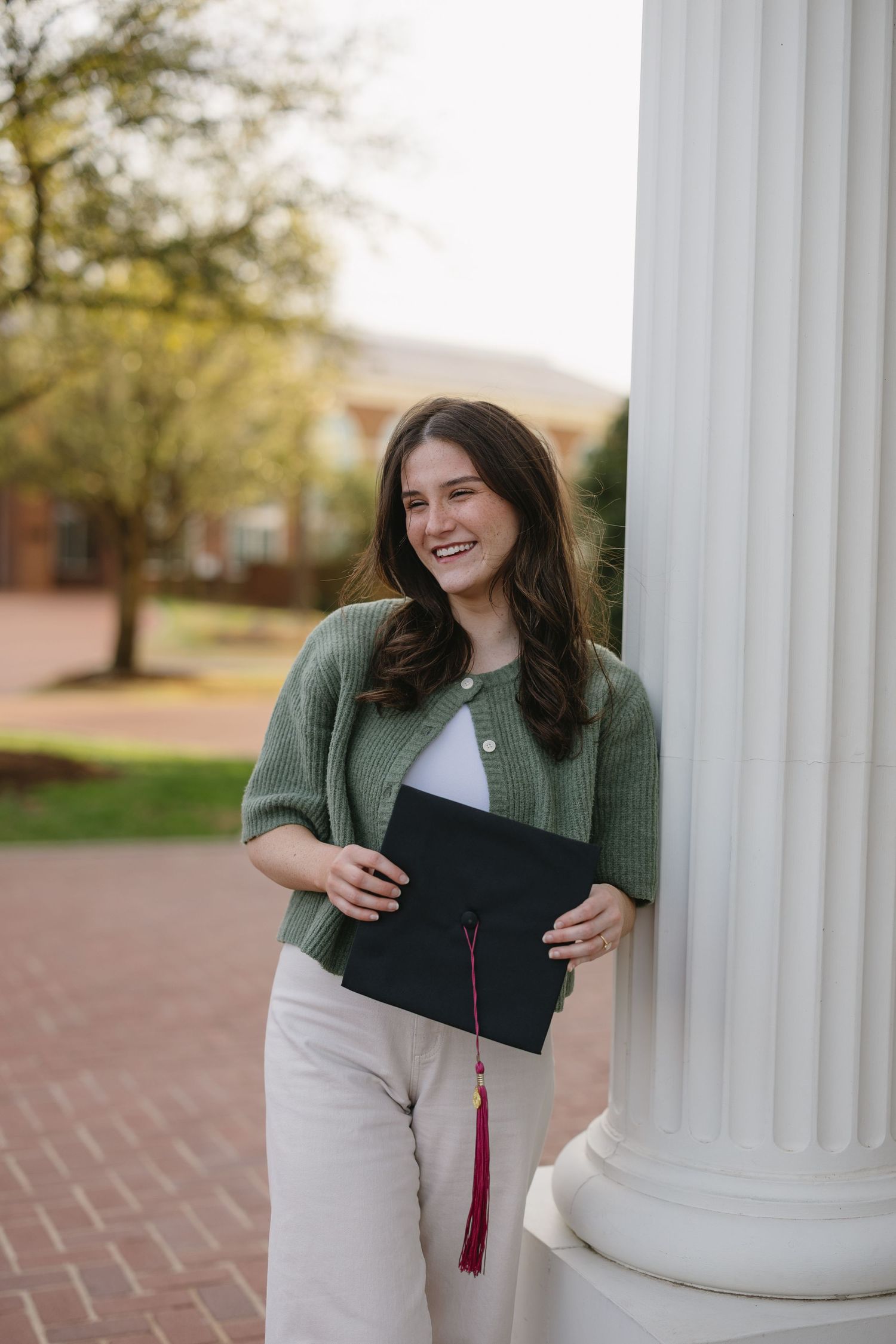 Smiling young woman in green cardigan holding graduation cap while leaning against a white column outdoors.