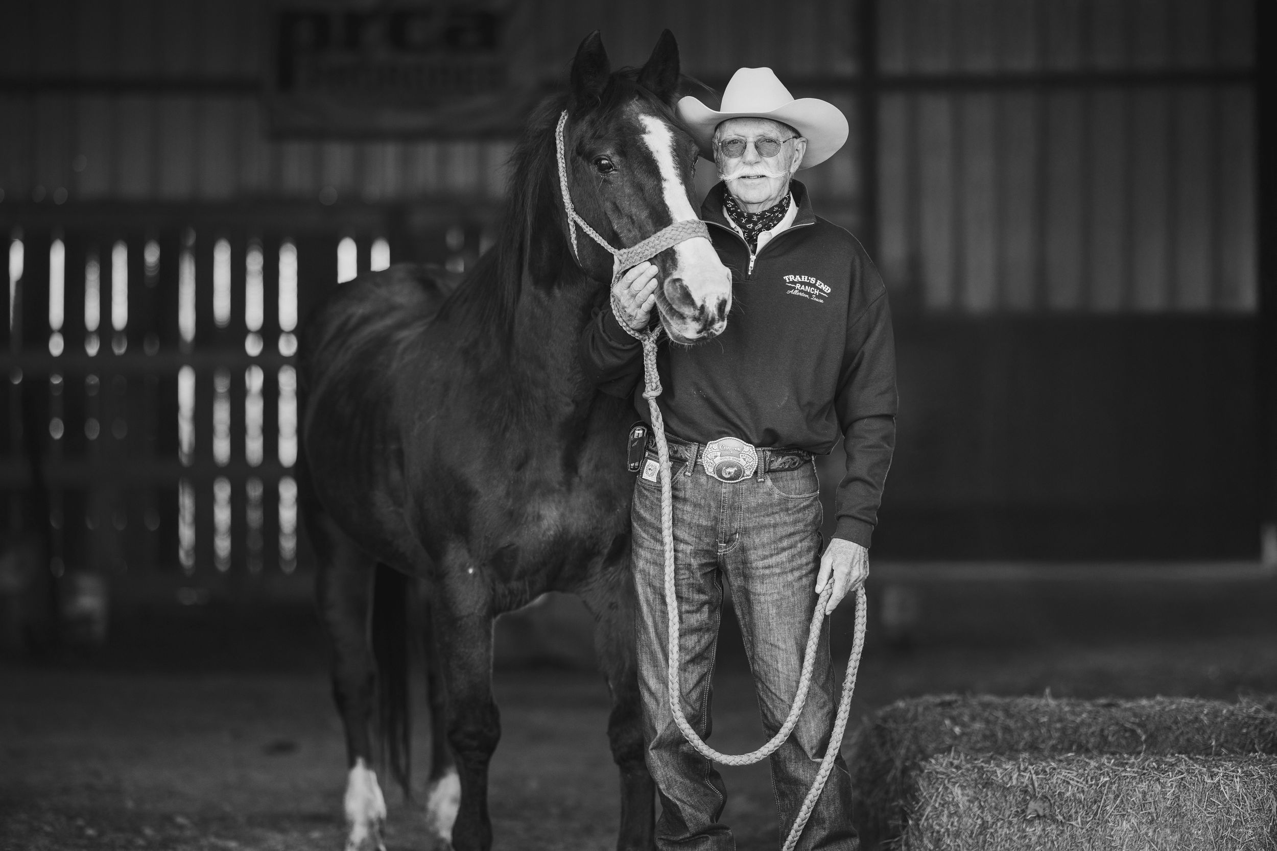 A black and white photograph shows a cowboy in a western hat leading a horse through a grassy pasture.