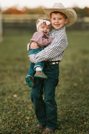 Two young children in cowboy hats and denim embrace while playing outdoors on grass in matching western outfits.