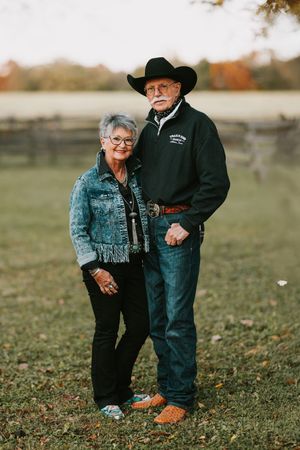 An elderly couple stands together outdoors wearing western attire with a lake and autumn trees in the background.