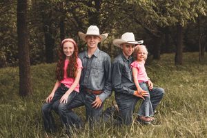 A rustic country family portrait of four people wearing cowboy hats and casual attire sitting together outdoors.