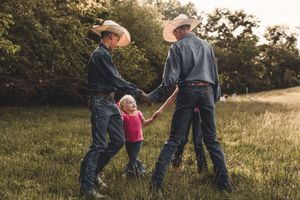 Three people in western wear walking together through a grassy field at sunset.