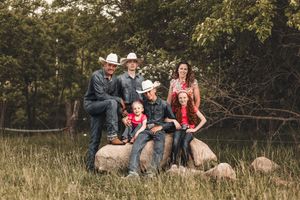 A group wearing cowboy hats and western attire poses together on large rocks in a grassy field with trees in background.