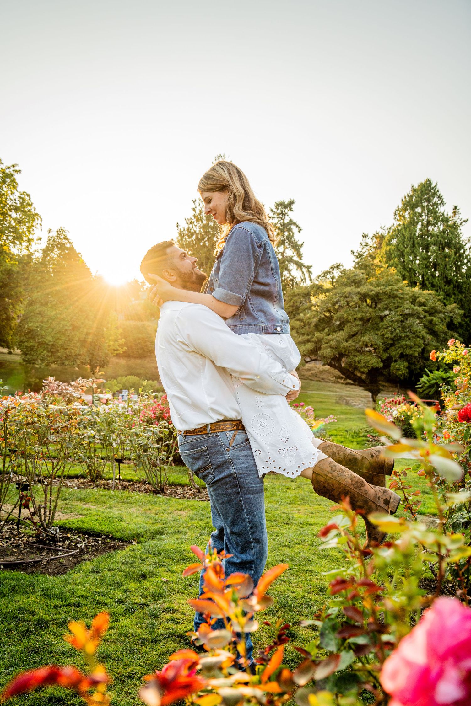 A couple embraces in a sunlit garden with colorful flowers in the foreground during golden hour.