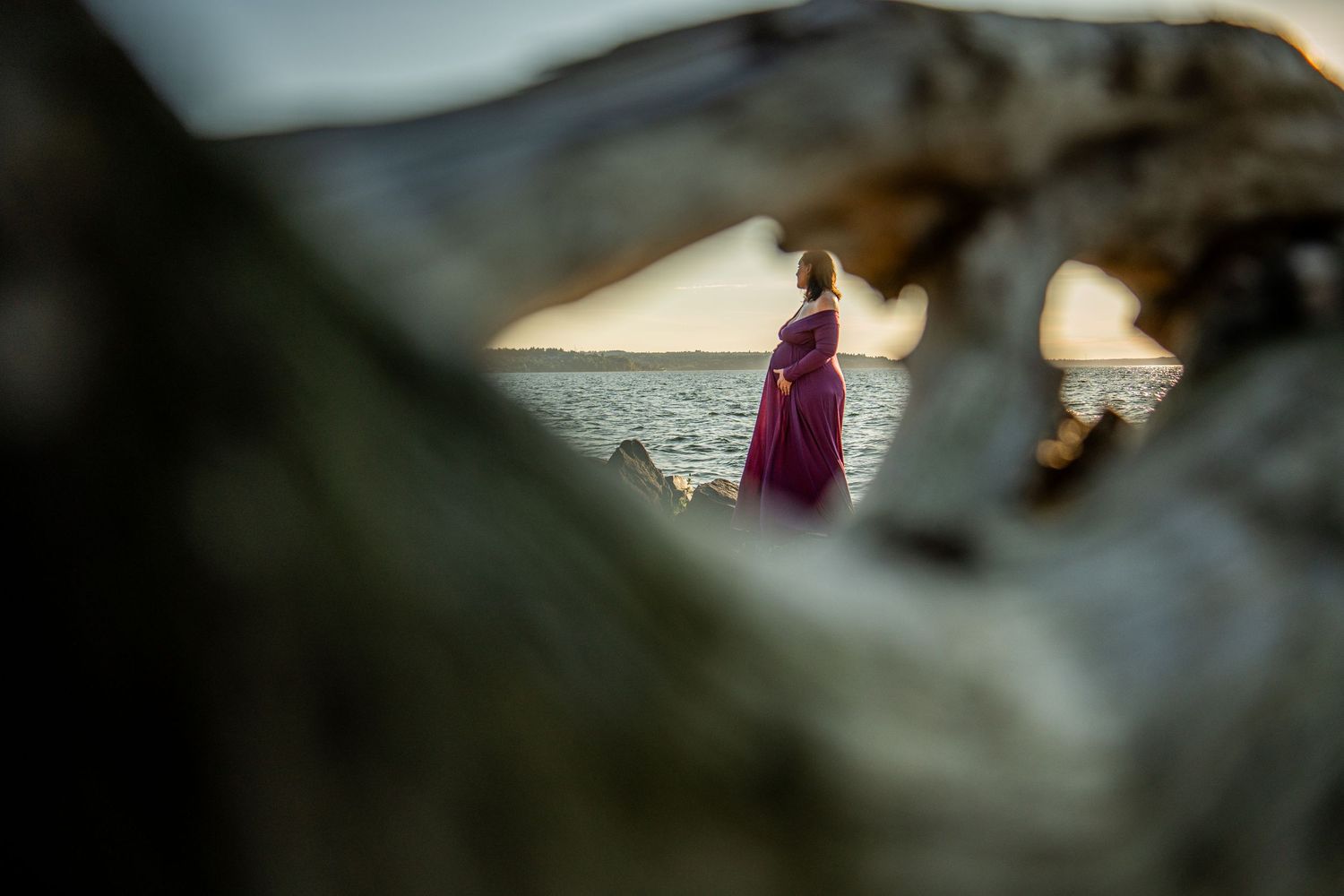 A person in a purple dress stands on a rocky beach at sunset with mountains in the background.