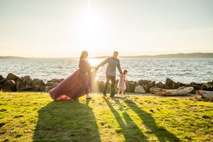 Two silhouettes hold hands on grassy shoreline at sunset with ocean and rocks in background.