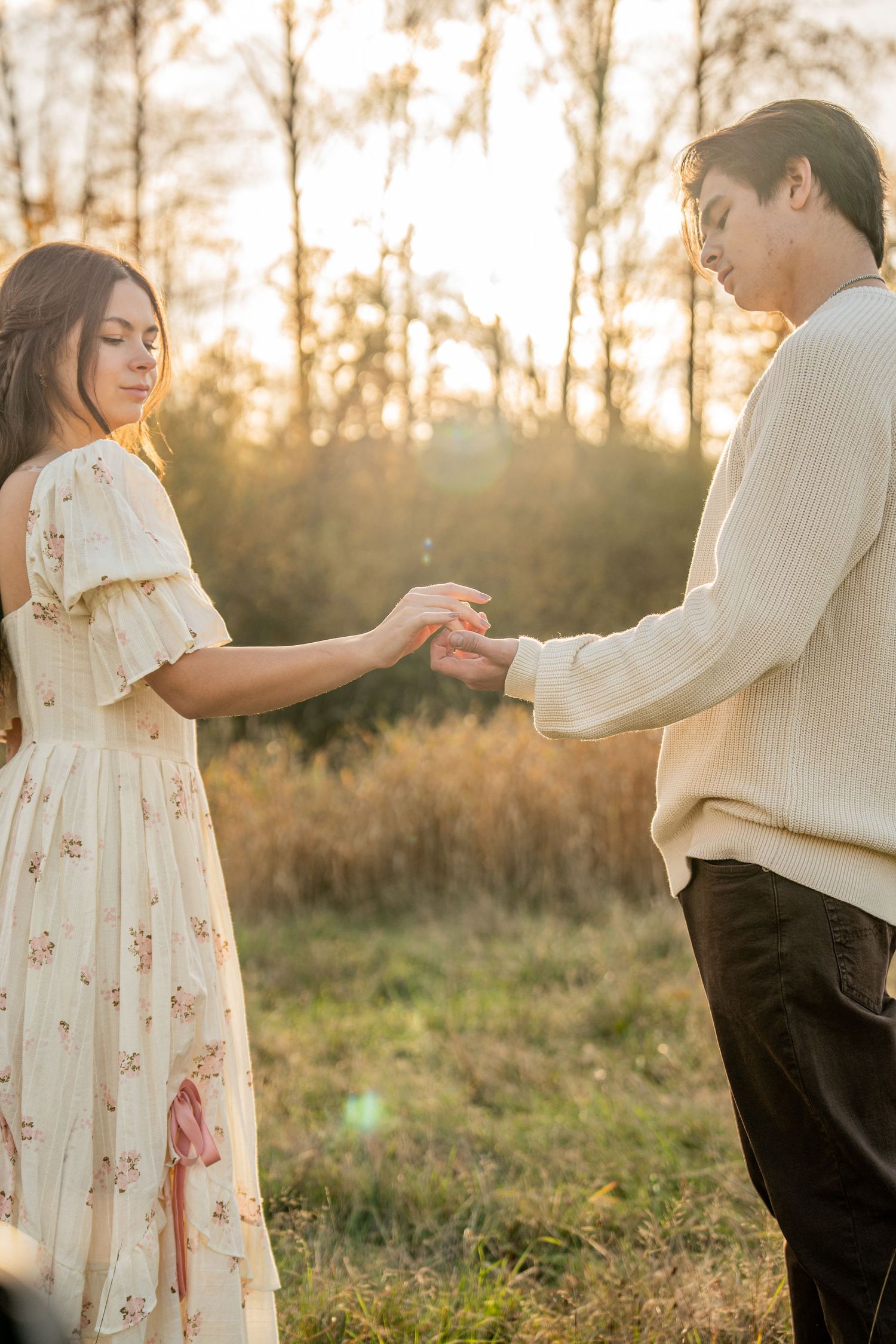Romantic couple in floral dress and casual attire share tender moments during golden hour sunset in a grassy field.