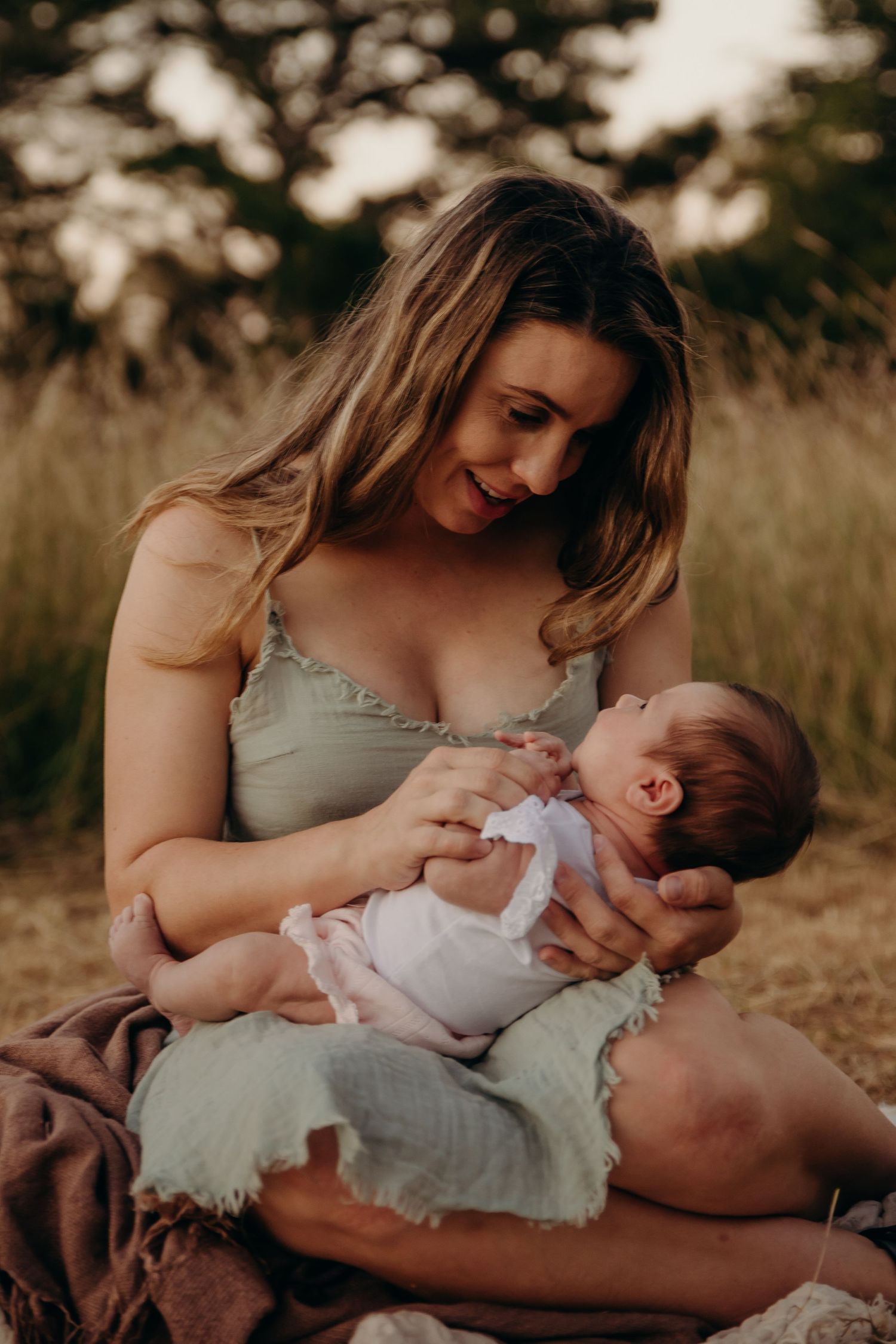 Mother smiles lovingly while cradling her newborn baby in a sunlit grassy outdoor setting.