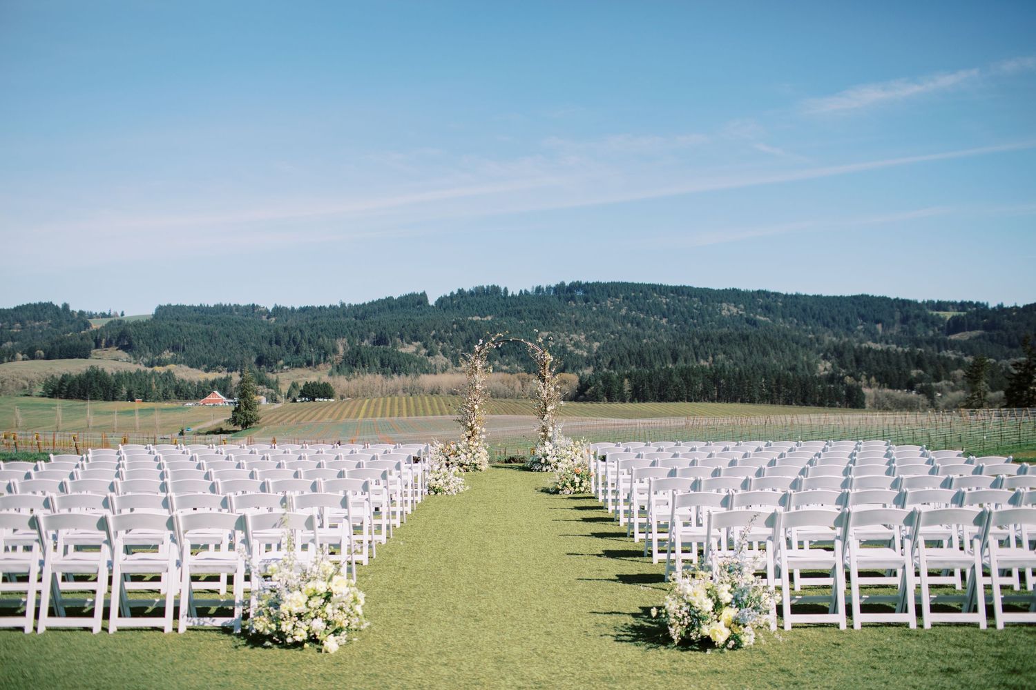 Rows of white folding chairs line a green lawn leading to a wooden wedding arch against rolling hills and blue sky.