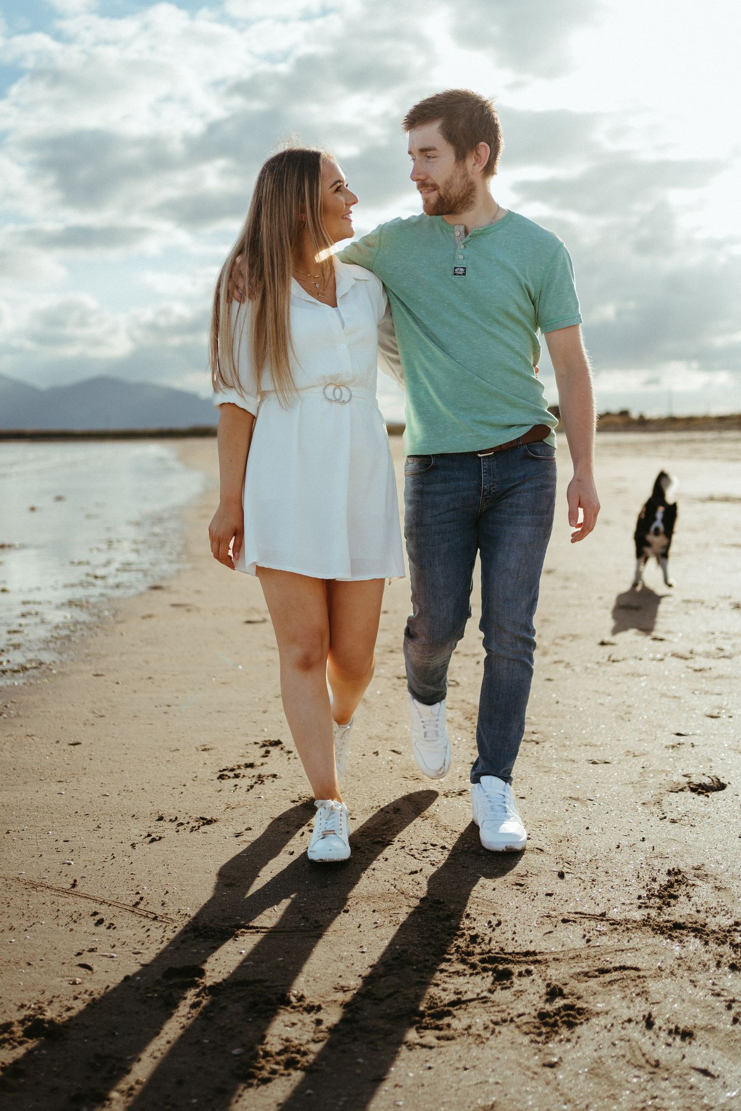 Couple walking along a beach shoreline with long shadows cast on the sand during sunset.