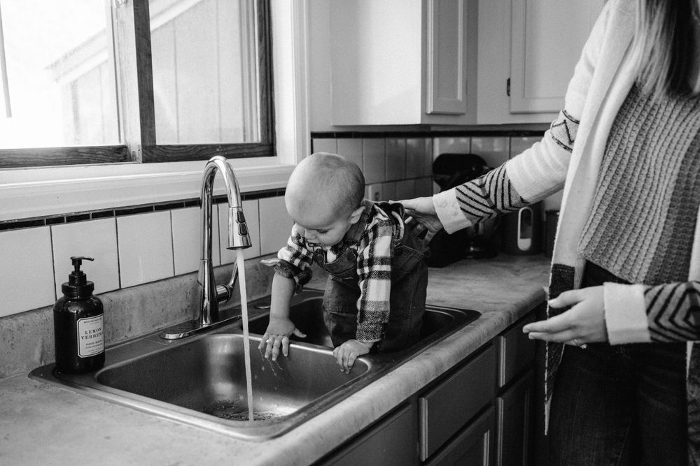 Child exploring kitchen sink while supervised in black and white photo.
