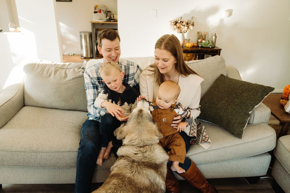 Family relaxing on gray couch with husky dog in sunny living room.
