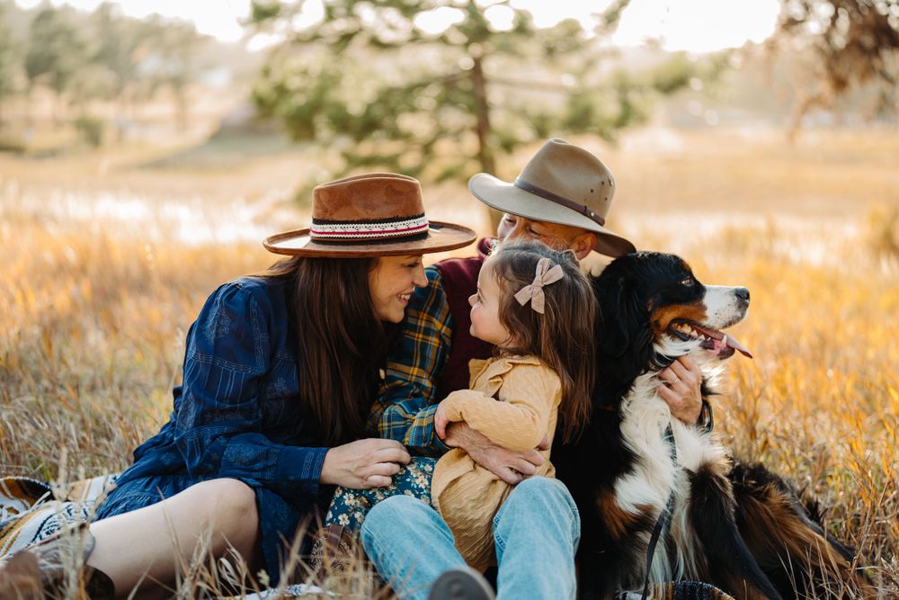 Two people in hats sit together on a blanket with their dog in a golden autumn field at sunset.