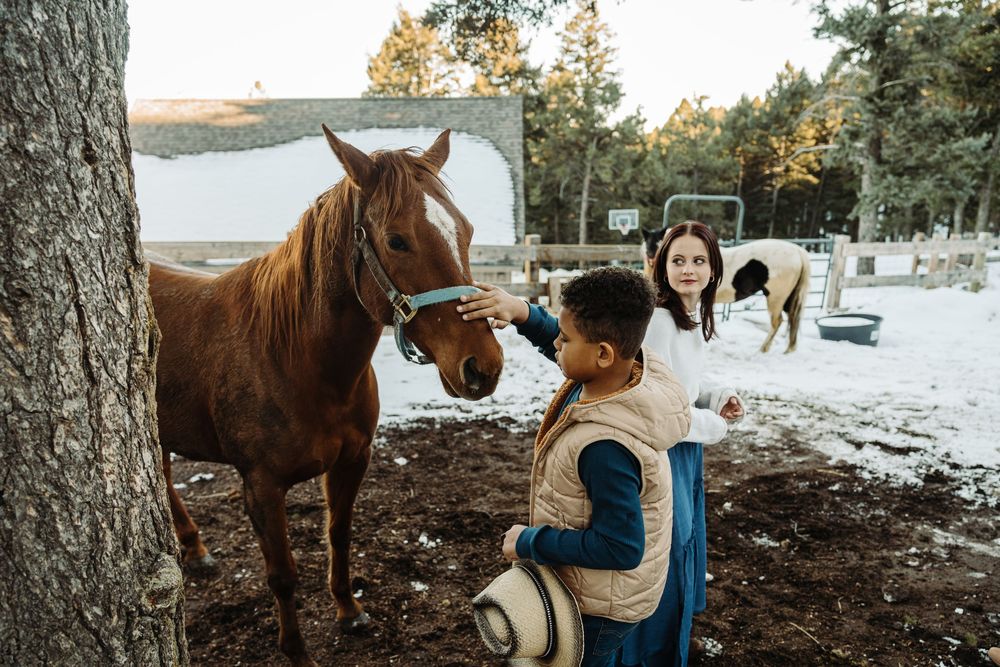 A woman and child petting a brown horse at a snowy farm with pine trees in the background.