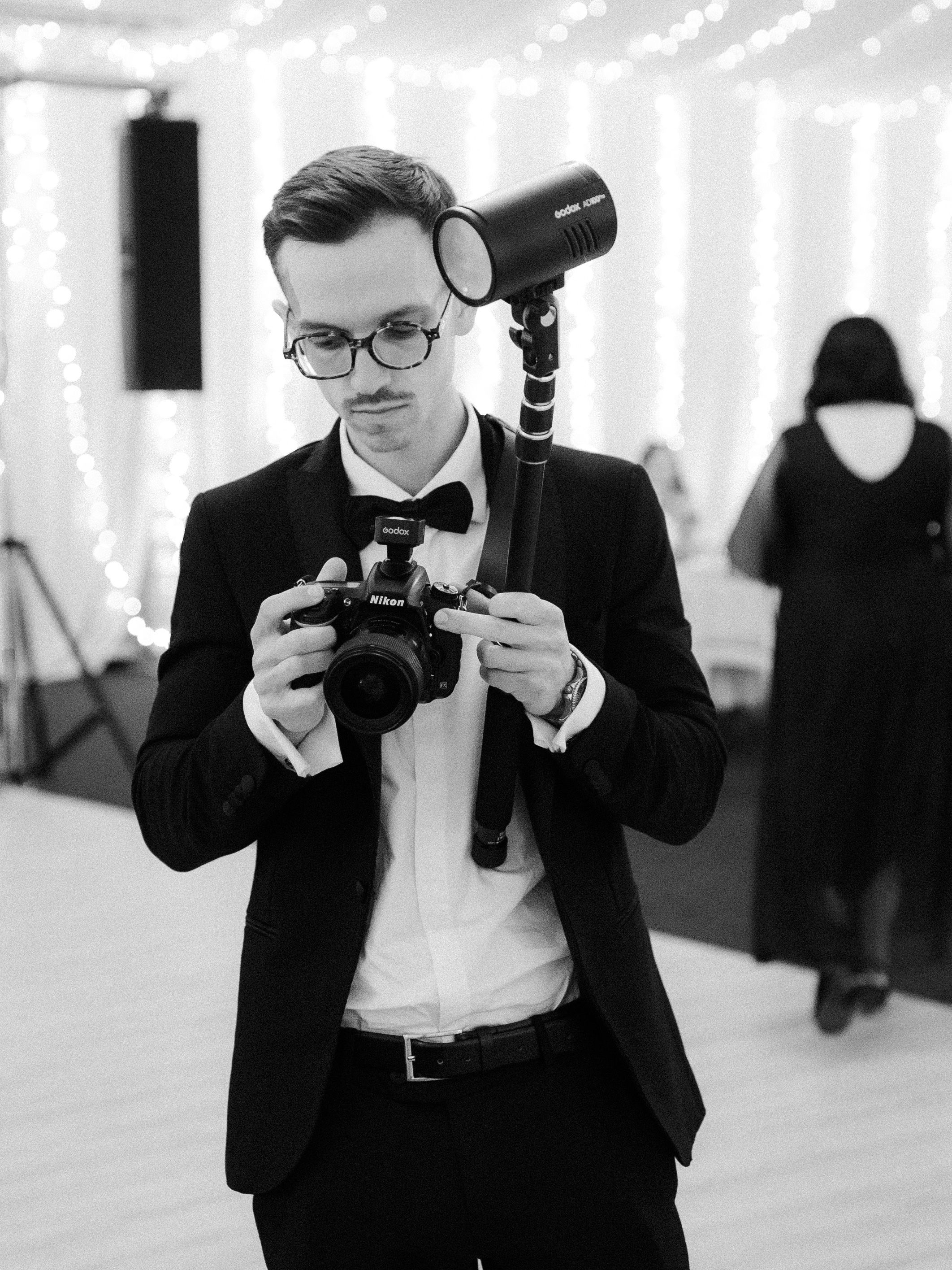 Professional photographer wearing formal attire and glasses examines camera at event in monochrome composition.