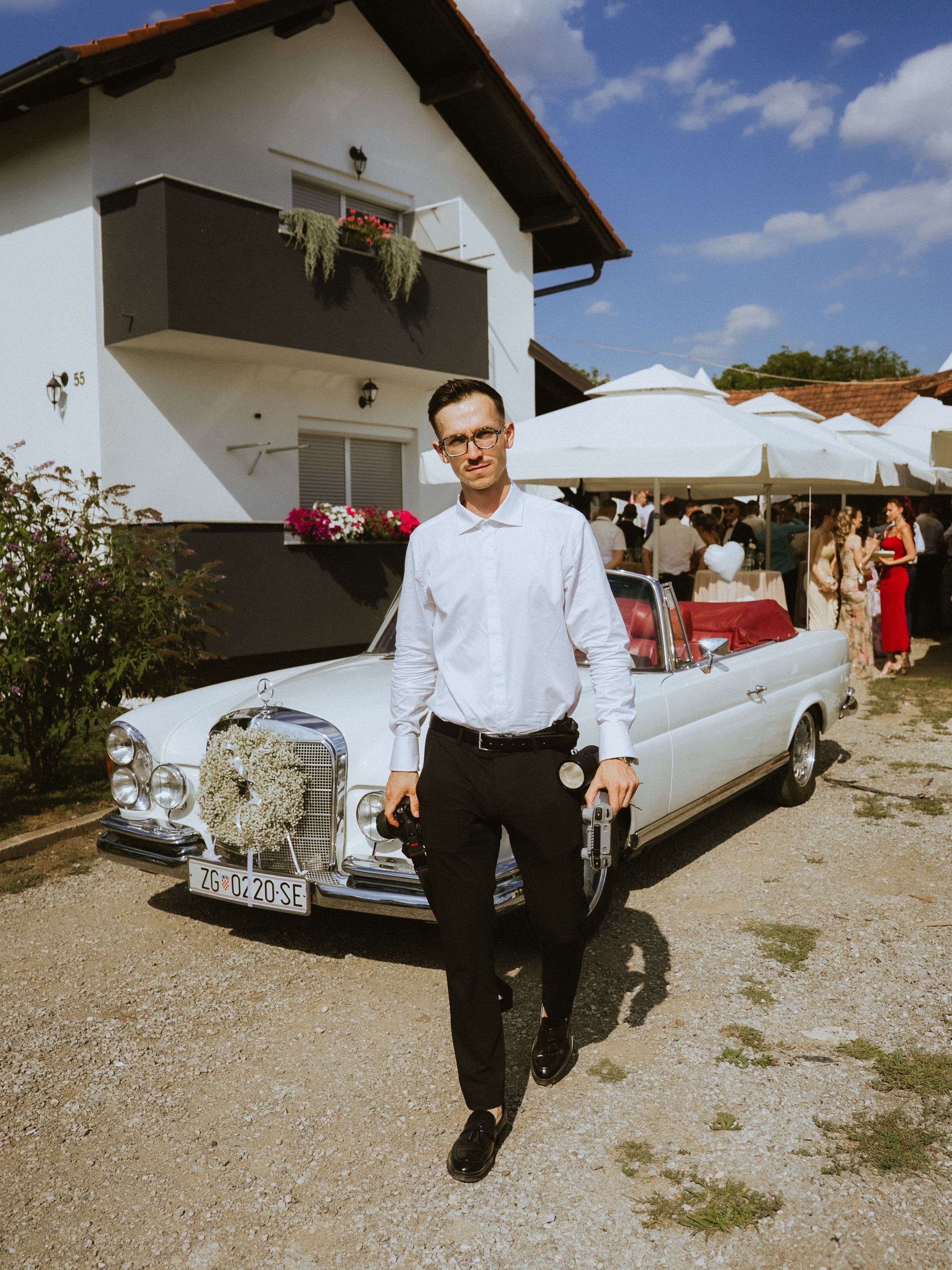 A vintage white Mercedes-Benz parked outside a house with flowers and patio umbrellas on a sunny day.