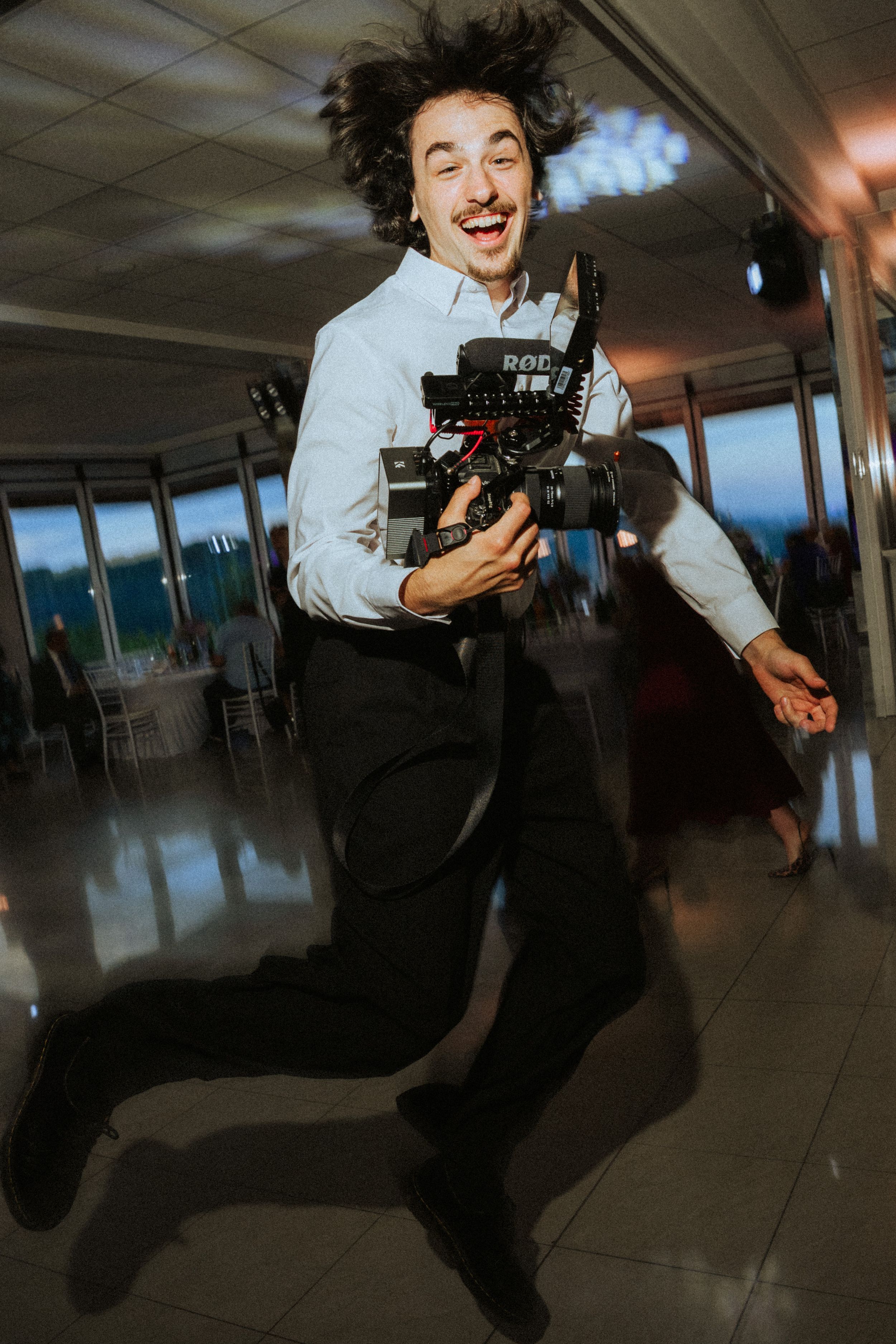 Excited videographer jumping mid-air holding a professional camera with RODEmic at an elegant indoor event venue.