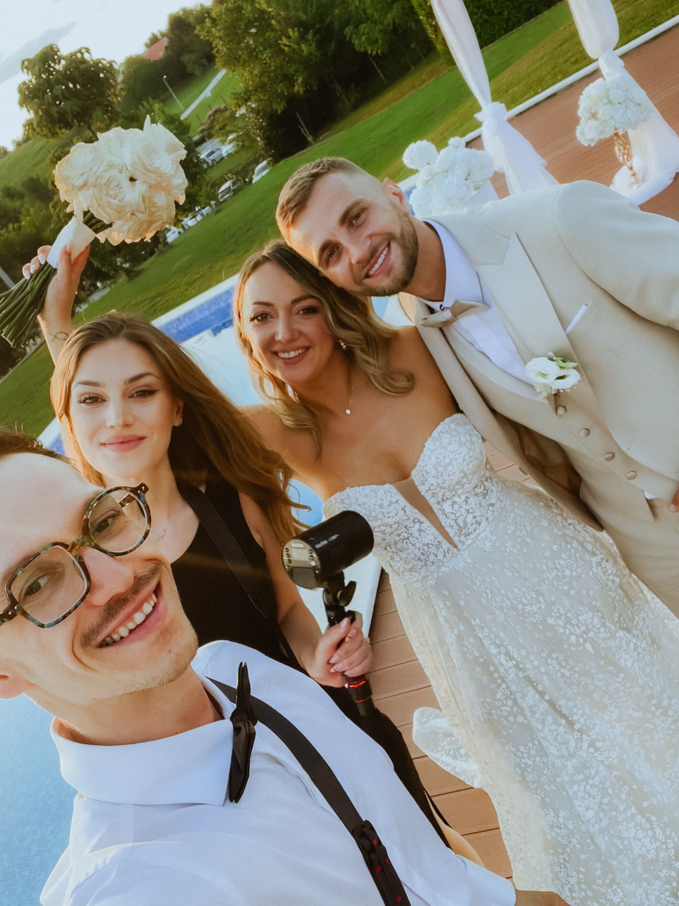 A joyful selfie moment captured outdoors at a wedding celebration set against a green lawn and summer sky.
