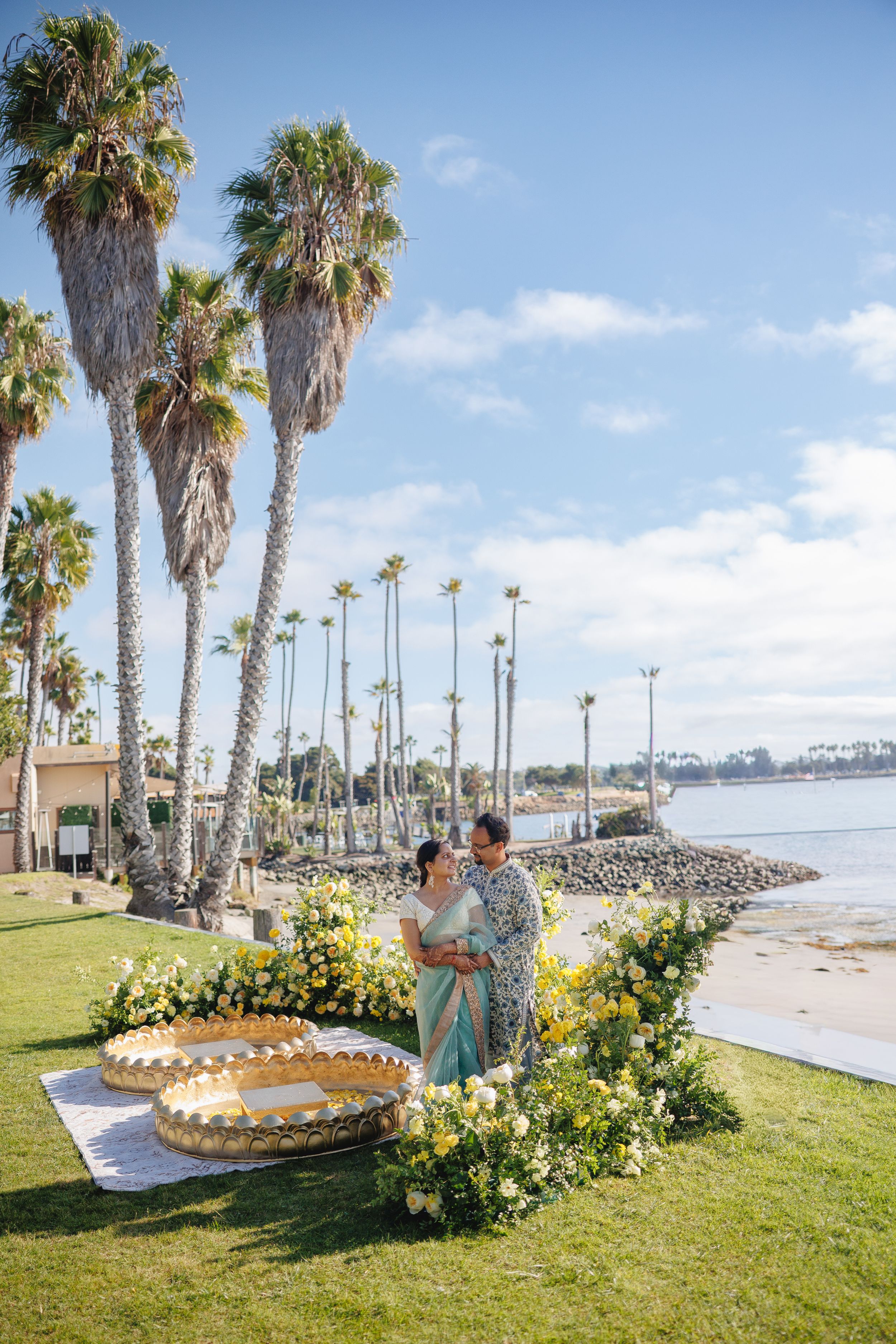 A coastal wedding ceremony setup with palm trees, yellow flowers, and a decorative seashell arch along the beach.
