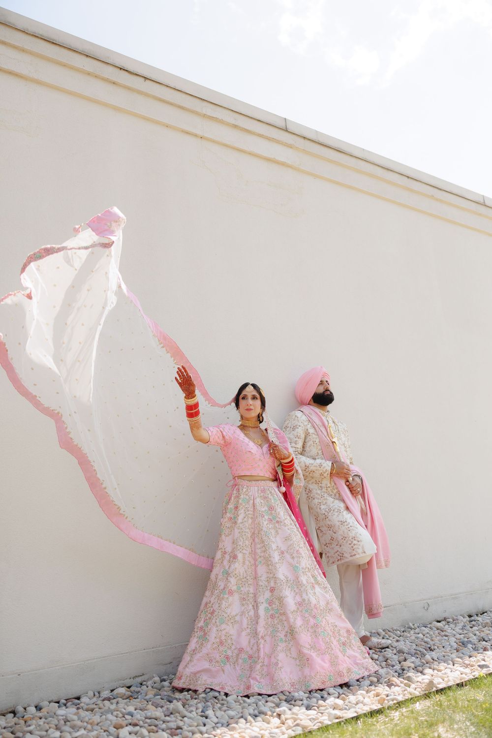Traditional Indian bride in pink and white lehenga poses gracefully with flowing dupatta against white wall.