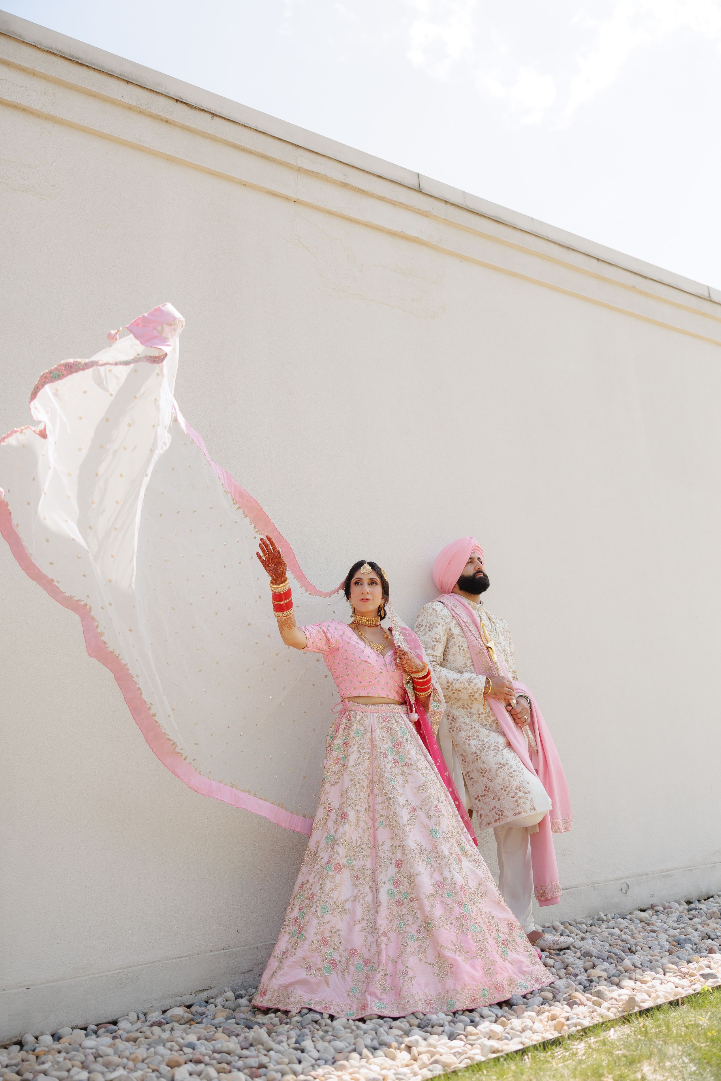 Traditional Indian bride in pink and white lehenga poses gracefully with flowing dupatta against white wall.