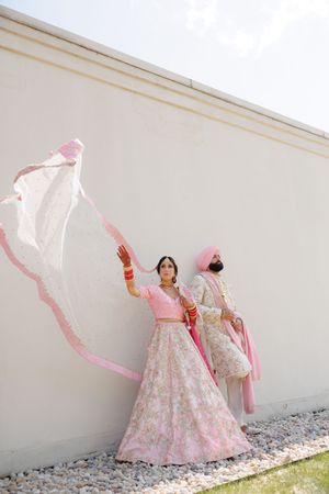 Traditional Indian bride in pink and white lehenga poses gracefully with flowing dupatta against white wall.