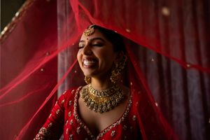 A traditional Indian bride wearing a red veil and ornate gold jewelry smiles during her wedding ceremony.