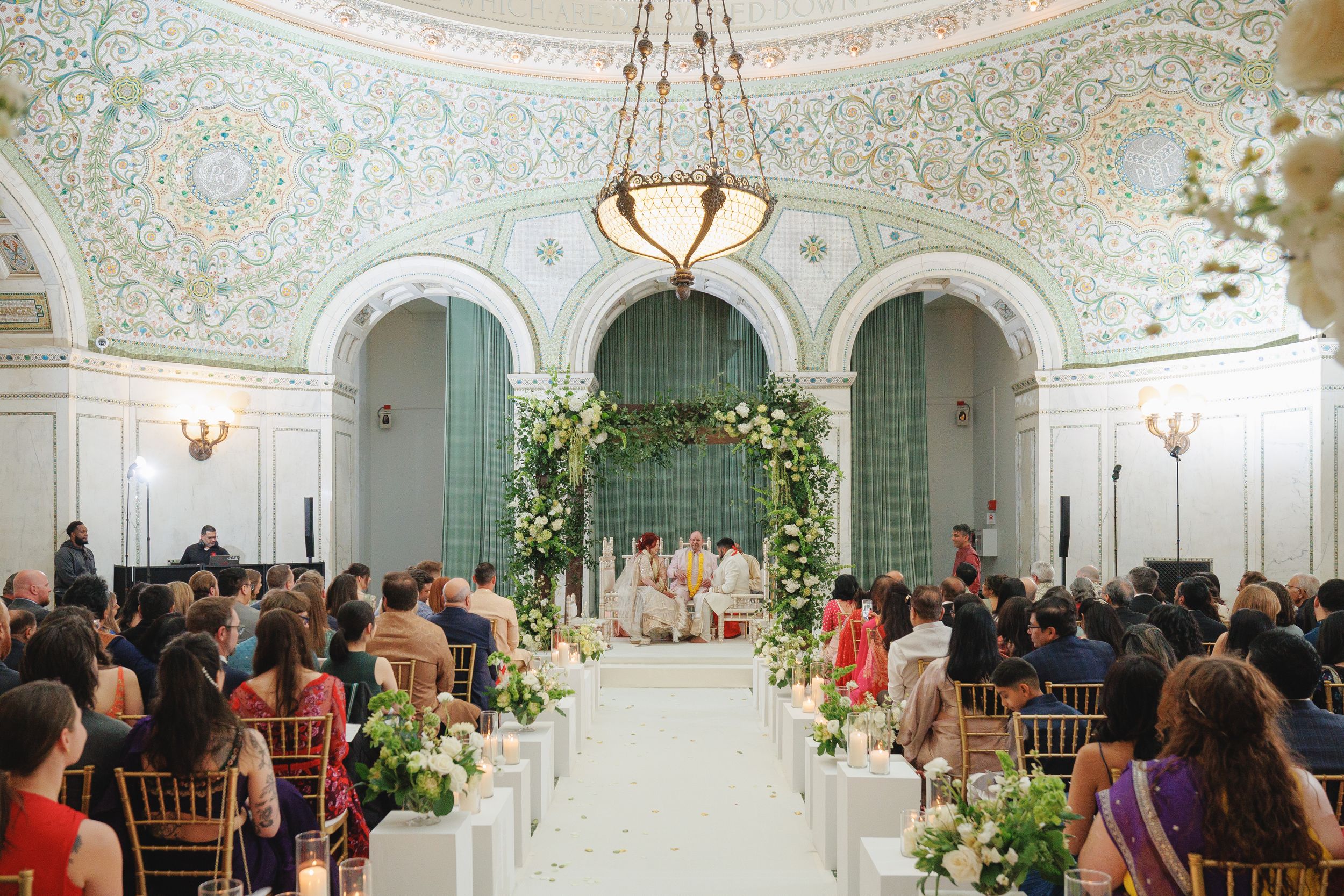 Elegant wedding ceremony in ornate ballroom with floral arch, white aisle, and gold chairs under decorative ceiling.