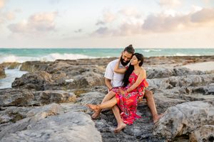 A couple embraces on rocky coastal shoreline during sunset with ocean waves and cloudy sky in background.