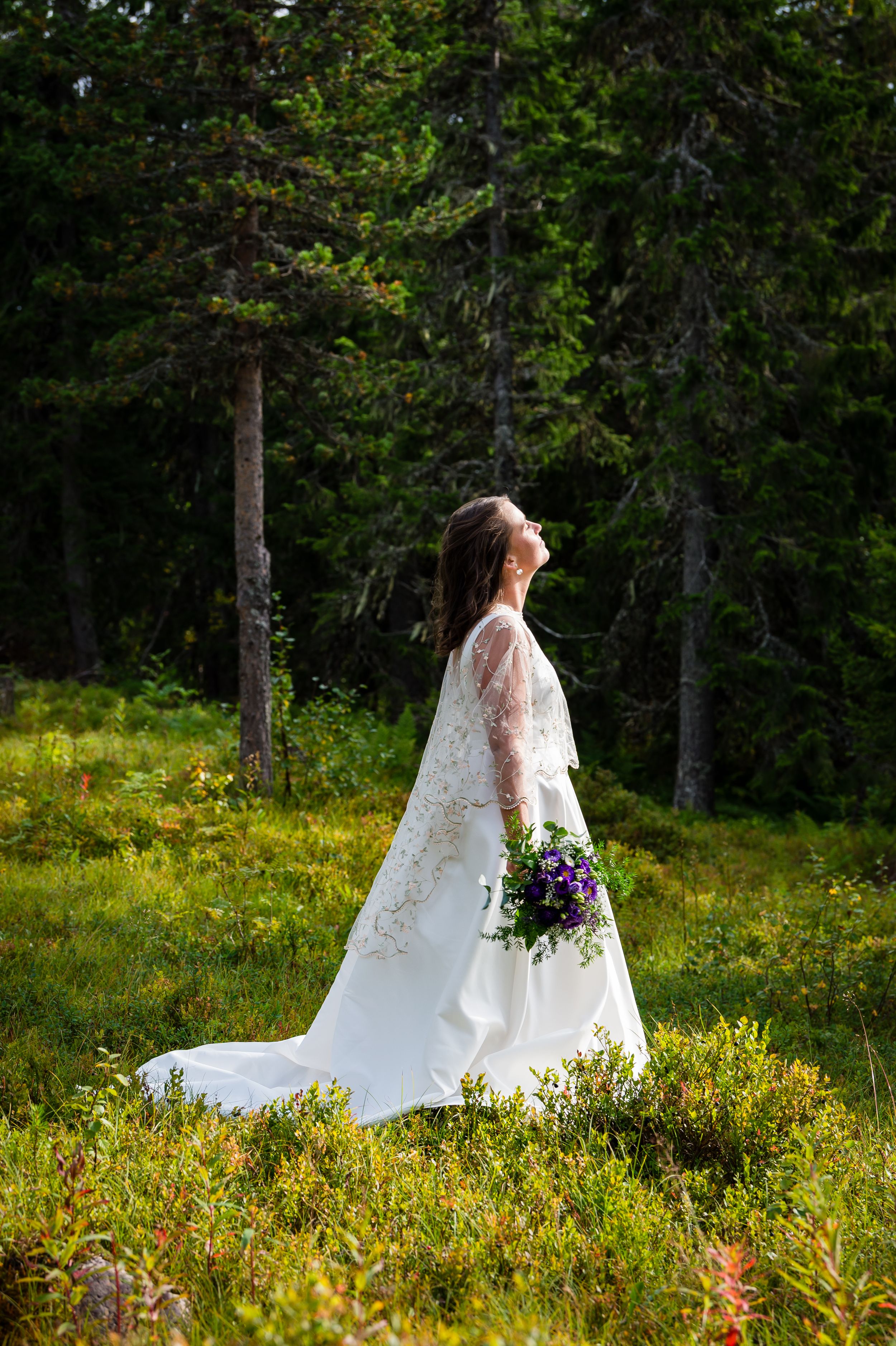 Wedding dress silhouette against forest backdrop with flowing veil and purple bouquet in sunlit meadow.